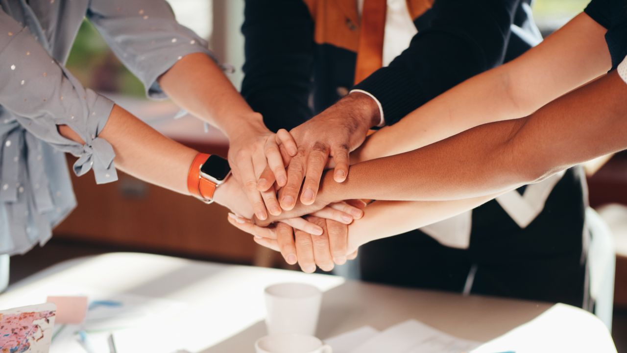 services-03 A close-up shot of diverse team members stacking hands over a white table, symbolizing unity and collaboration.
